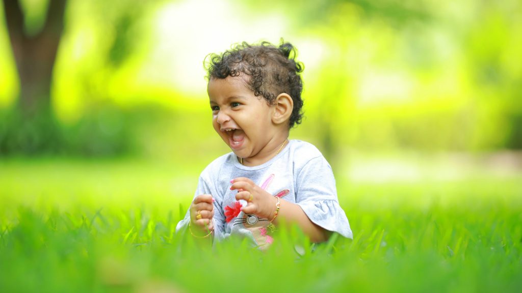 Toddler laughing while sitting on grass outdoors, representing the importance of outdoor play in early childhood development.