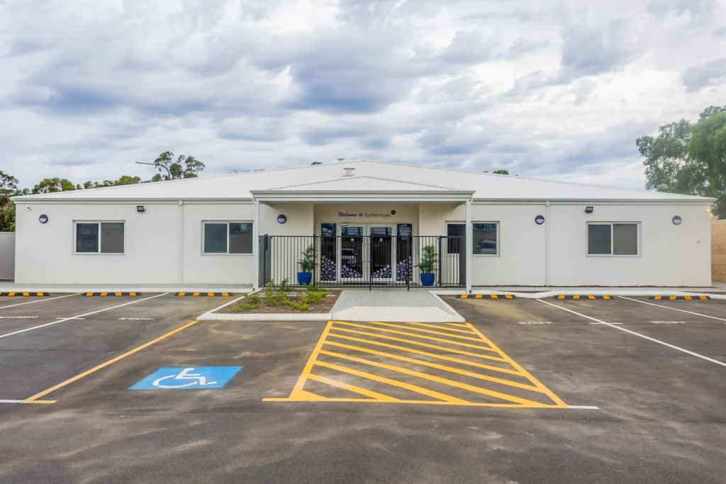 The front entrance of Buttercups Perth Childcare, a modern white building with a paved parking area featuring clearly marked accessibility spaces. The entrance is framed by a small garden and a secure black gate, providing a welcoming and accessible environment for families and children.