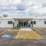 The front entrance of Buttercups Perth Childcare, a modern white building with a paved parking area featuring clearly marked accessibility spaces. The entrance is framed by a small garden and a secure black gate, providing a welcoming and accessible environment for families and children.