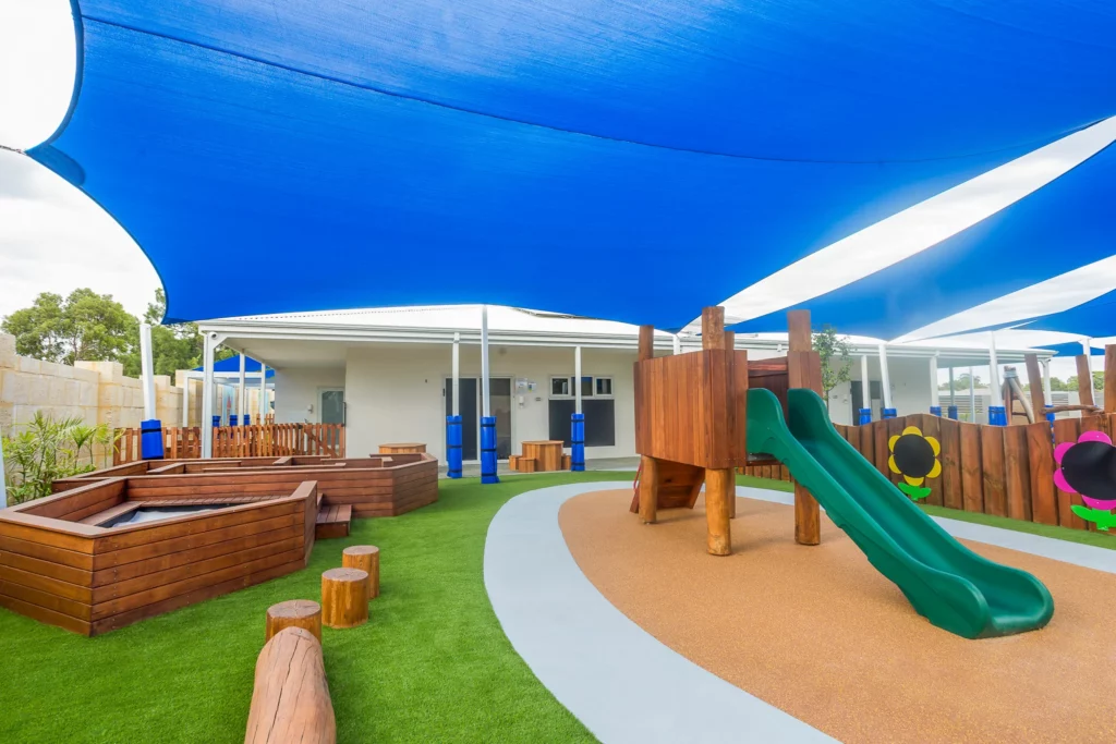 A vibrant outdoor play area at Buttercups Perth Childcare featuring a wooden structure with a green slide, surrounded by artificial grass and natural wood stepping logs. Shaded by expansive blue canopies, the space includes a sandpit and colourful flower-shaped boards on the wooden fence, creating a stimulating and safe environment for children to explore and play.