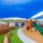 A vibrant outdoor play area at Buttercups Perth Childcare featuring a wooden structure with a green slide, surrounded by artificial grass and natural wood stepping logs. Shaded by expansive blue canopies, the space includes a sandpit and colourful flower-shaped boards on the wooden fence, creating a stimulating and safe environment for children to explore and play.