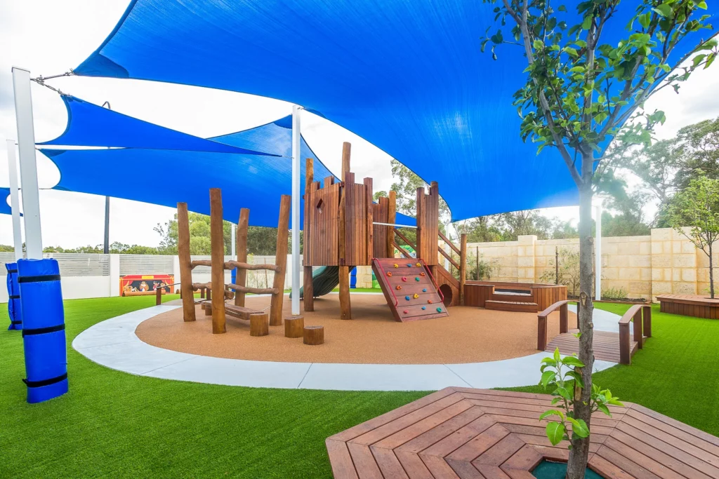 A dynamic outdoor play area at Buttercups Perth Childcare showcasing a wooden climbing structure with a colourful rock-climbing wall and rope bridges. The space is shaded with large blue sail canopies, surrounded by green artificial turf and natural wooden elements, creating a fun and safe environment for children.