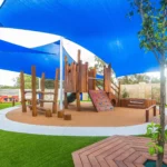 A dynamic outdoor play area at Buttercups Perth Childcare showcasing a wooden climbing structure with a colourful rock-climbing wall and rope bridges. The space is shaded with large blue sail canopies, surrounded by green artificial turf and natural wooden elements, creating a fun and safe environment for children.