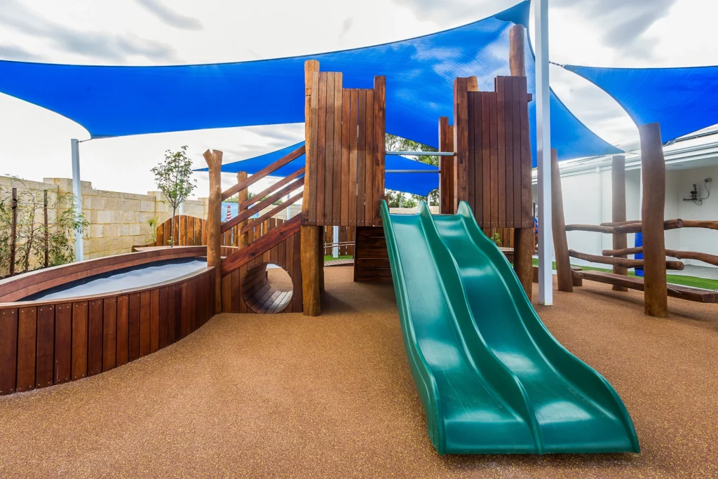 A dynamic outdoor play structure at Buttercups Perth Childcare featuring a double green slide, wooden climbing ramps, and a tunnel entrance, all set on a soft, rubberised surface and shaded by blue sail canopies, providing a safe and engaging environment for children to explore and play.