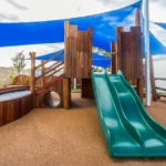 A dynamic outdoor play structure at Buttercups Perth Childcare featuring a double green slide, wooden climbing ramps, and a tunnel entrance, all set on a soft, rubberised surface and shaded by blue sail canopies, providing a safe and engaging environment for children to explore and play.