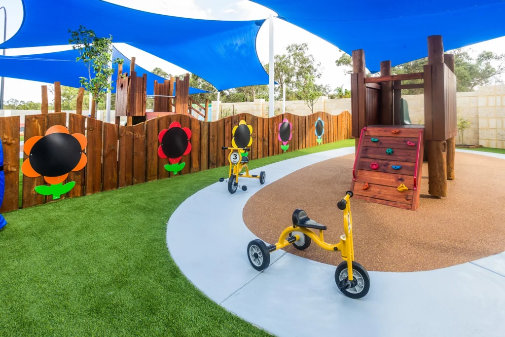 A vibrant outdoor play area at Buttercups Perth Childcare featuring yellow tricycles parked on a concrete path, a wooden climbing wall with colourful handholds, and decorative flower-shaped chalkboards on a wooden fence, all shaded by bright blue sail canopies, creating a fun and safe environment for children.