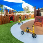 A vibrant outdoor play area at Buttercups Perth Childcare featuring yellow tricycles parked on a concrete path, a wooden climbing wall with colourful handholds, and decorative flower-shaped chalkboards on a wooden fence, all shaded by bright blue sail canopies, creating a fun and safe environment for children.