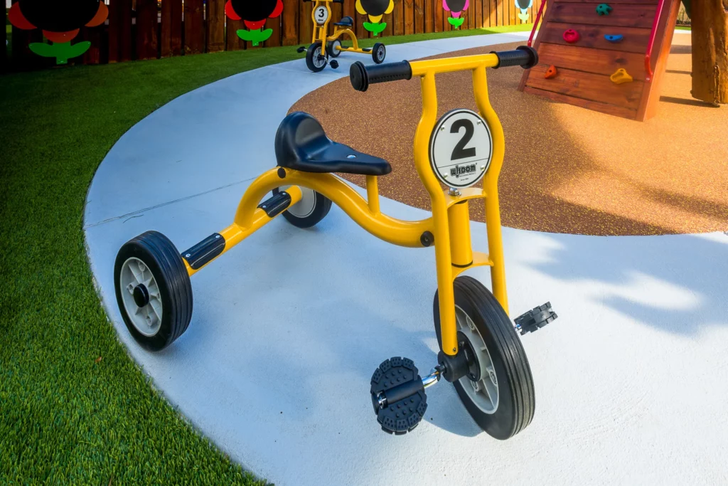 A bright yellow tricycle labelled with the number "2," parked on a clean concrete pathway in the outdoor play area at Buttercups Perth Childcare, surrounded by green artificial turf and a climbing wall in the background, offering children an engaging and active play experience.