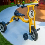 A bright yellow tricycle labelled with the number "2," parked on a clean concrete pathway in the outdoor play area at Buttercups Perth Childcare, surrounded by green artificial turf and a climbing wall in the background, offering children an engaging and active play experience.