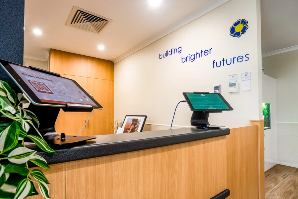 The welcoming reception area at Buttercups Perth Childcare, featuring modern check-in kiosks on a wooden counter with a digital display, greenery, and the phrase "building brighter futures" prominently displayed on the wall, reflecting the centre's commitment to quality childcare.