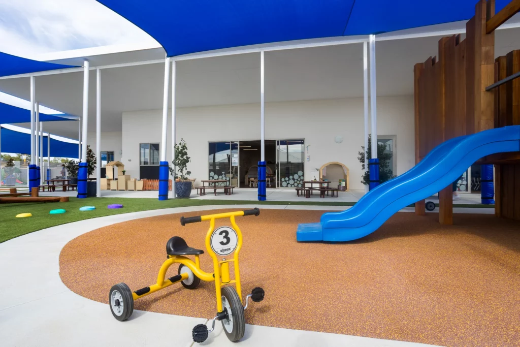 A well-designed outdoor play area at Buttercups Perth Childcare, featuring a yellow tricycle on a soft rubber surface near a vibrant blue slide. The area includes shaded structures, grassy play zones, and access to indoor learning spaces, offering a safe and stimulating environment for children.