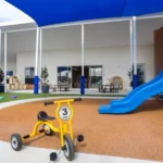 A well-designed outdoor play area at Buttercups Perth Childcare, featuring a yellow tricycle on a soft rubber surface near a vibrant blue slide. The area includes shaded structures, grassy play zones, and access to indoor learning spaces, offering a safe and stimulating environment for children.