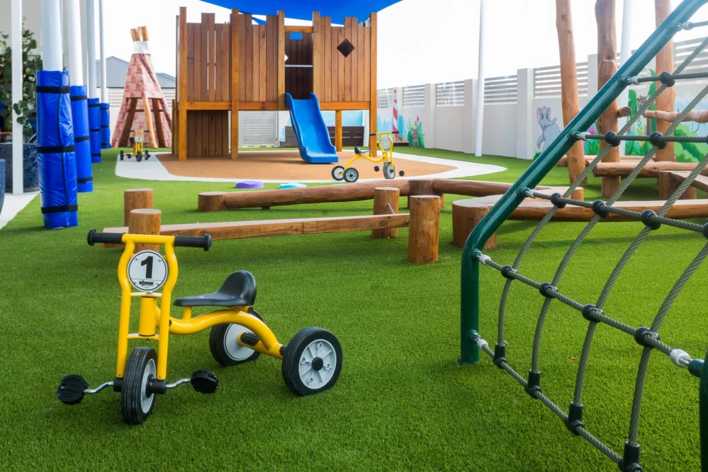 A vibrant outdoor play area at Buttercups Perth Childcare featuring a wooden fort with a blue slide, a balance beam, and a climbing net. The grassy space includes a yellow tricycle, wooden play structures, and shaded areas, fostering a fun and engaging environment for children to explore and enjoy.