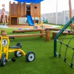 A vibrant outdoor play area at Buttercups Perth Childcare featuring a wooden fort with a blue slide, a balance beam, and a climbing net. The grassy space includes a yellow tricycle, wooden play structures, and shaded areas, fostering a fun and engaging environment for children to explore and enjoy.