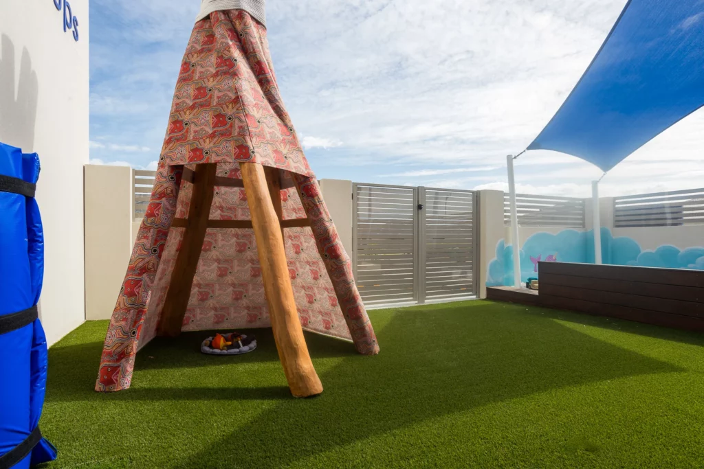 A teepee-style structure covered in a vibrant, patterned fabric, set on a grassy play area at Buttercups Perth Childcare. The outdoor space features shaded seating, fencing, and a mural of clouds and playful designs, creating a welcoming and creative environment for children.