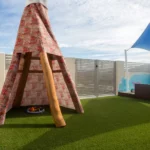 A teepee-style structure covered in a vibrant, patterned fabric, set on a grassy play area at Buttercups Perth Childcare. The outdoor space features shaded seating, fencing, and a mural of clouds and playful designs, creating a welcoming and creative environment for children.