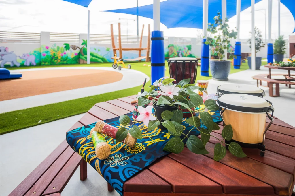 A colourful outdoor play area at Buttercups Perth Childcare featuring a wooden table adorned with traditional drums, maracas, flowers, and cultural decor. The background includes vibrant murals, play equipment, and shaded seating areas for creative and interactive learning.