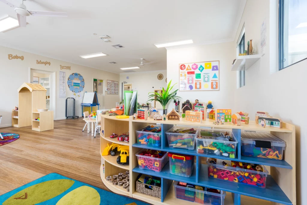 A vibrant childcare classroom featuring organised shelves filled with colourful toys, blocks, and educational materials. The room includes a shape recognition poster, natural light, and interactive play areas, promoting learning and creativity at Buttercups Perth Childcare.