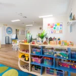 A vibrant childcare classroom featuring organised shelves filled with colourful toys, blocks, and educational materials. The room includes a shape recognition poster, natural light, and interactive play areas, promoting learning and creativity at Buttercups Perth Childcare.