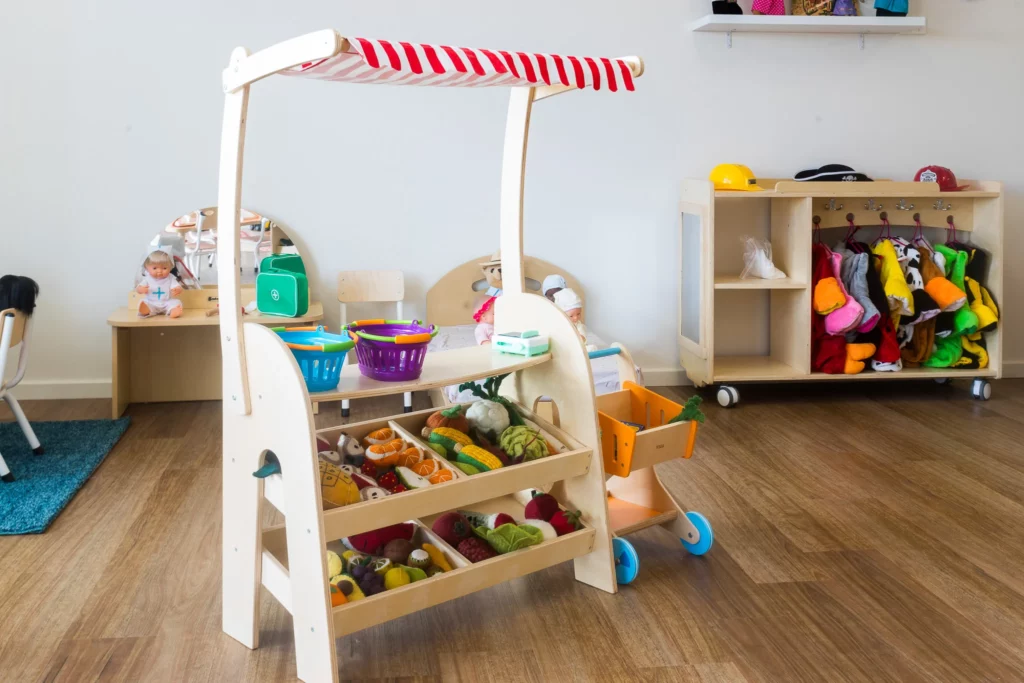 Role-playing corner at Buttercups Perth Childcare with a wooden market stand stocked with toy fruits and vegetables, dress-up costumes, and a doll care station, encouraging imaginative play and creativity.