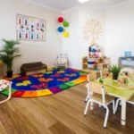 Interactive play area at Buttercups Perth Childcare featuring a colourful alphabet rug, wooden tables with toys, bookshelves, and vibrant decor, creating a fun and nurturing environment for children.