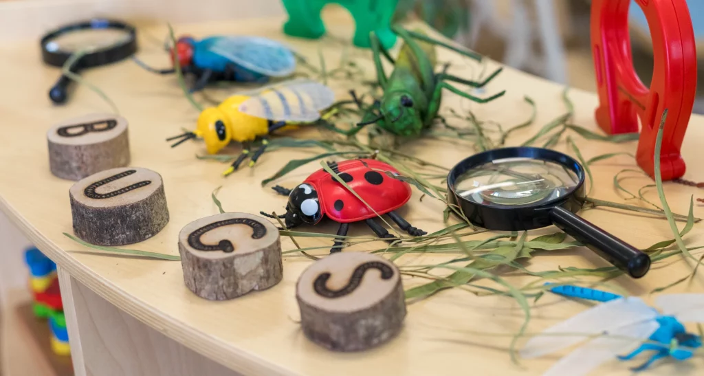 Close-up view of a nature exploration table setup at Buttercups Perth Childcare. The table features colourful toy insects, a magnifying glass, and wooden discs with 'BUGS' spelled out, encouraging imaginative play and curiosity in children.