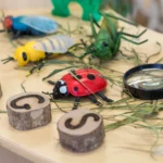 Close-up view of a nature exploration table setup at Buttercups Perth Childcare. The table features colourful toy insects, a magnifying glass, and wooden discs with 'BUGS' spelled out, encouraging imaginative play and curiosity in children.