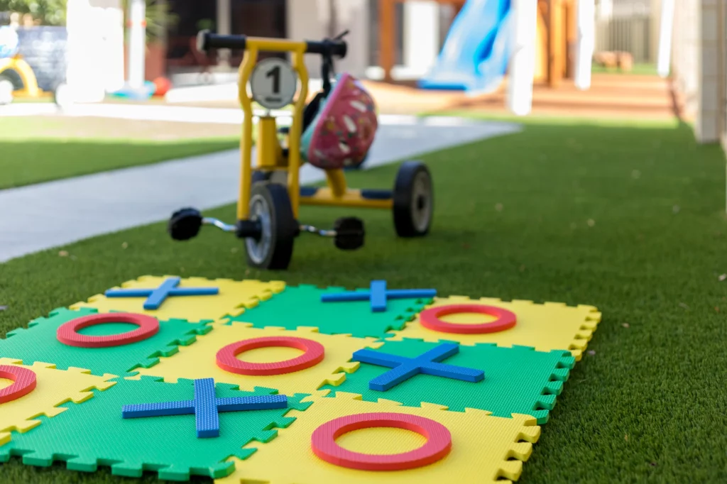 Close-up of the outdoor play area at Buttercups Perth Childcare featuring a tic-tac-toe foam game mat and a tricycle on artificial grass, promoting active and creative play.