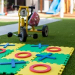 Close-up of the outdoor play area at Buttercups Perth Childcare featuring a tic-tac-toe foam game mat and a tricycle on artificial grass, promoting active and creative play.