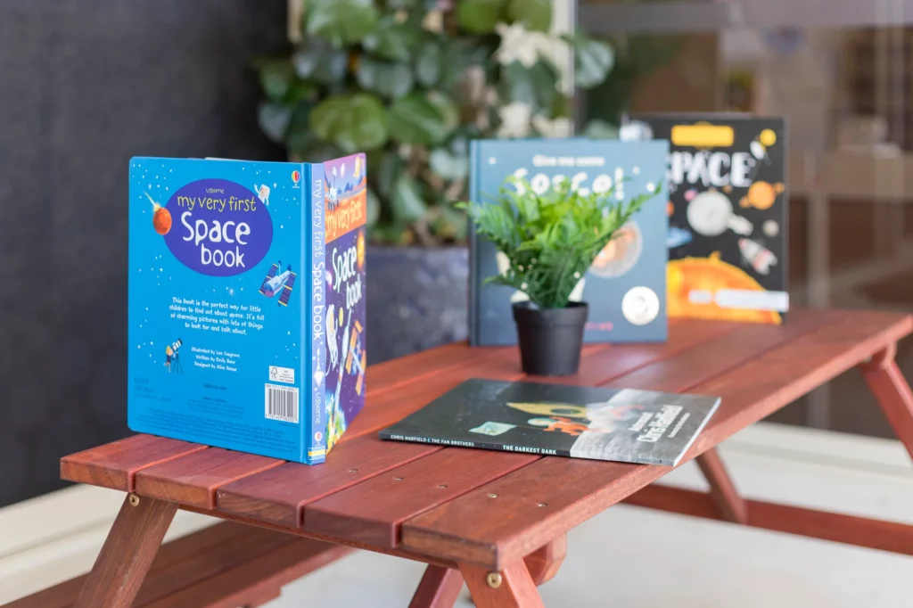 A collection of children's space-themed books, including "My Very First Space Book," displayed on a small wooden table at Buttercups Perth Childcare. A small green potted plant adds a touch of greenery to the setup, creating a welcoming learning environment.