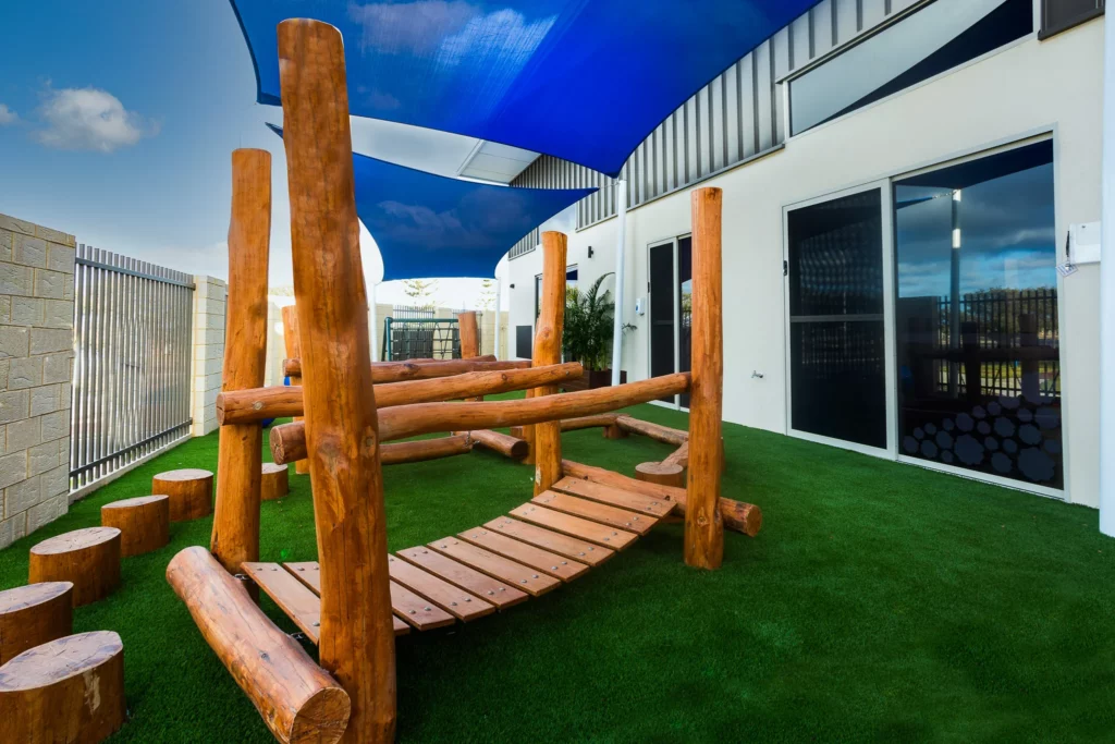 Natural outdoor play area at Buttercups Perth Childcare featuring wooden climbing structures, soft artificial grass, and shaded spaces, designed to encourage exploration, balance, and active play in a safe environment.