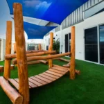 Natural outdoor play area at Buttercups Perth Childcare featuring wooden climbing structures, soft artificial grass, and shaded spaces, designed to encourage exploration, balance, and active play in a safe environment.