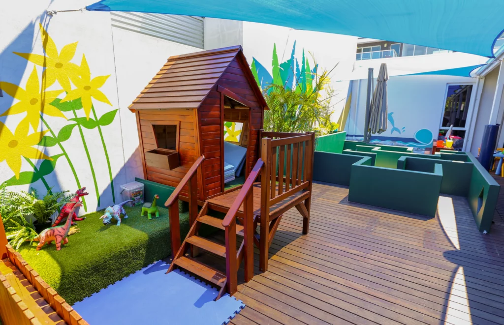 Vibrant outdoor play area at a Perth childcare centre, featuring a wooden cubby house, toy dinosaurs, and creative activity spaces under shaded coverings.