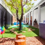 Natural outdoor play area at a Perth childcare centre, featuring tree stumps, shade from trees, and interactive activities for exploration and creativity.