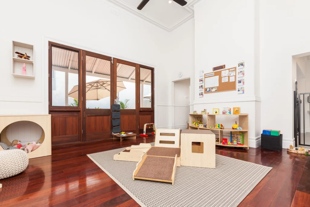 Bright and inviting childcare room at Buttercups Perth Childcare, featuring wooden flooring, a cosy play area with soft furnishings, a climbing structure, and neatly organised shelves filled with educational toys and books.