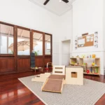 Bright and inviting childcare room at Buttercups Perth Childcare, featuring wooden flooring, a cosy play area with soft furnishings, a climbing structure, and neatly organised shelves filled with educational toys and books.