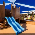 The wooden play structure with twin blue slides at Buttercups Perth Childcare is shaded by blue and white sails, offering a safe and fun environment for children.