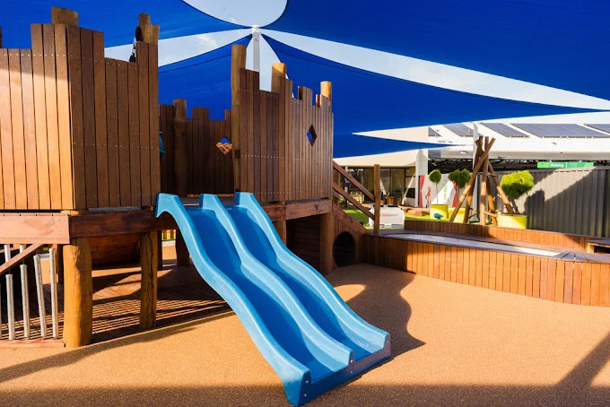 The wooden play structure with twin blue slides at Buttercups Perth Childcare is shaded by blue and white sails, offering a safe and fun environment for children.