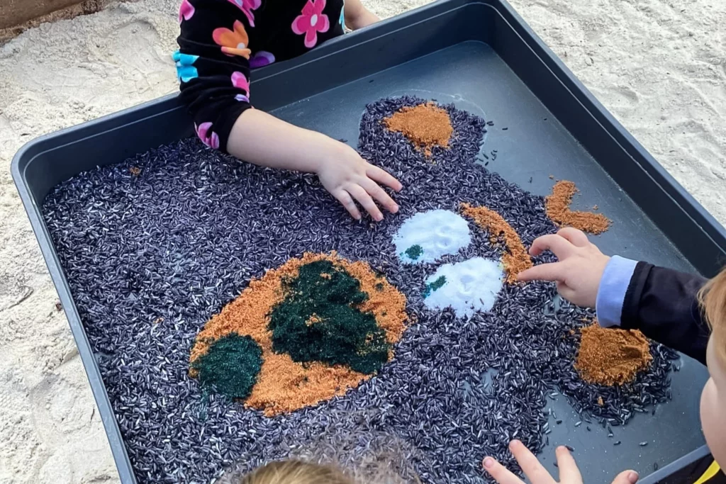 Children at Buttercups Perth Childcare engage in a sensory play activity, creating a colourful artwork in a tray filled with dyed rice and textured materials, fostering creativity and fine motor skills development.