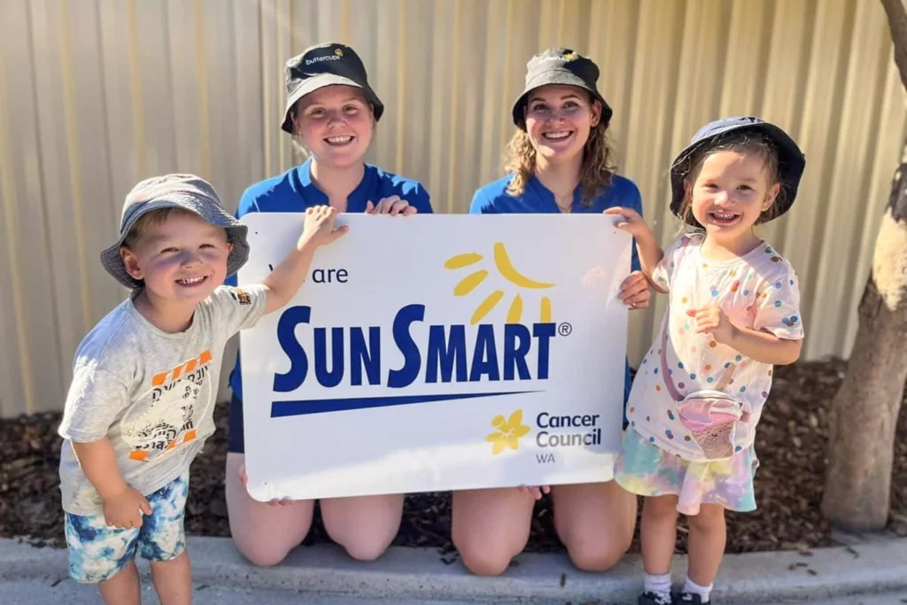 Two smiling educators from Buttercups Perth Childcare, wearing SunSmart hats, pose alongside two cheerful children holding a large "We are SunSmart" sign from Cancer Council WA, emphasising the centre's commitment to sun safety and wellbeing.