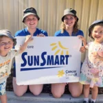 Two smiling educators from Buttercups Perth Childcare, wearing SunSmart hats, pose alongside two cheerful children holding a large "We are SunSmart" sign from Cancer Council WA, emphasising the centre's commitment to sun safety and wellbeing.