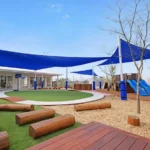 The outdoor area at Buttercups Perth Childcare showcases natural wooden log seating, a circular green play zone, climbing equipment, and slides under expansive blue shade sails.