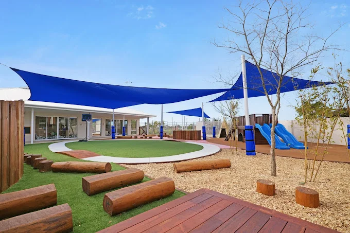 The outdoor area at Buttercups Perth Childcare showcases natural wooden log seating, a circular green play zone, climbing equipment, and slides under expansive blue shade sails.