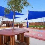 Outdoor learning and play space at Buttercups Perth Childcare featuring a wooden seating area, hopscotch markings, sand play zone, and climbing structures under protective blue shade sails.