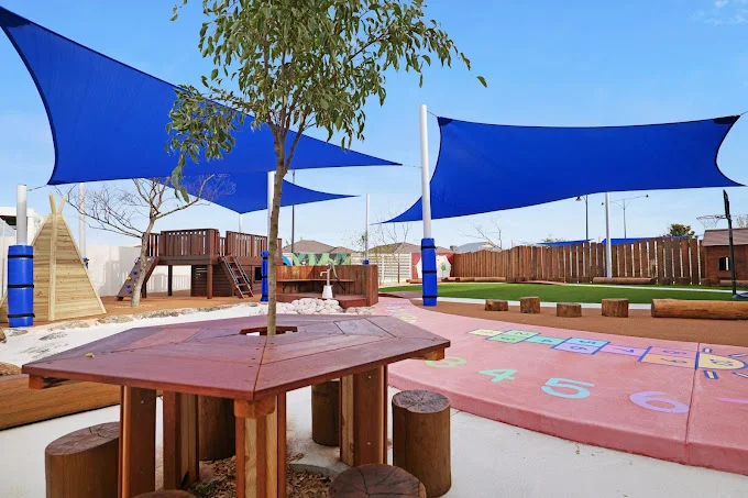 Outdoor learning and play space at Buttercups Perth Childcare featuring a wooden seating area, hopscotch markings, sand play zone, and climbing structures under protective blue shade sails.