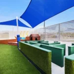 Outdoor activity area at Buttercups Perth Childcare features a green maze structure, shaded play spaces with blue sails, and vibrant playhouses designed for interactive learning and fun.