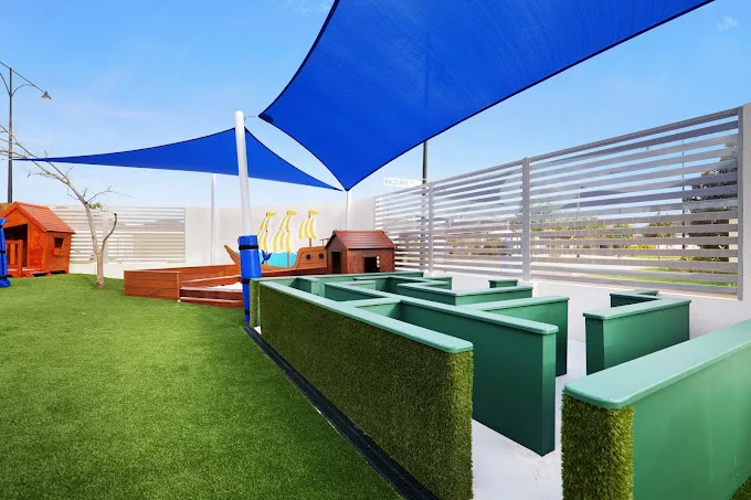 Outdoor activity area at Buttercups Perth Childcare features a green maze structure, shaded play spaces with blue sails, and vibrant playhouses designed for interactive learning and fun.