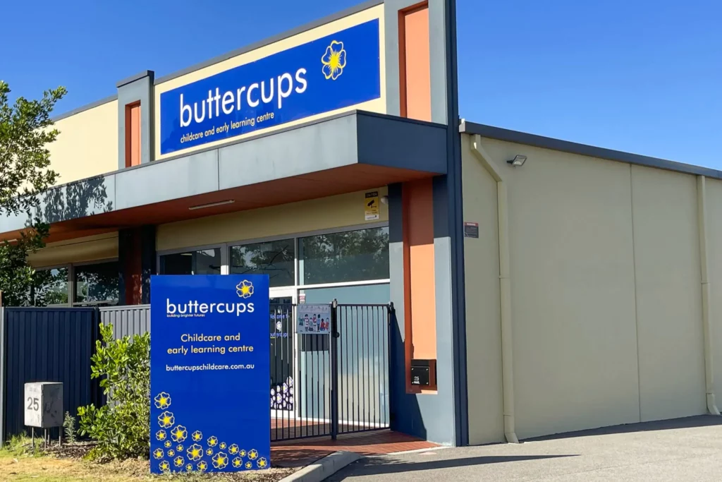 Exterior view of Buttercups Perth Childcare centre with a vibrant blue sign displaying the logo and text, "Childcare and early learning centre." The modern facility features a gated entrance and surrounding greenery under a clear blue sky, presenting a welcoming and professional environment.