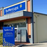 Exterior view of Buttercups Perth Childcare centre with a vibrant blue sign displaying the logo and text, "Childcare and early learning centre." The modern facility features a gated entrance and surrounding greenery under a clear blue sky, presenting a welcoming and professional environment.