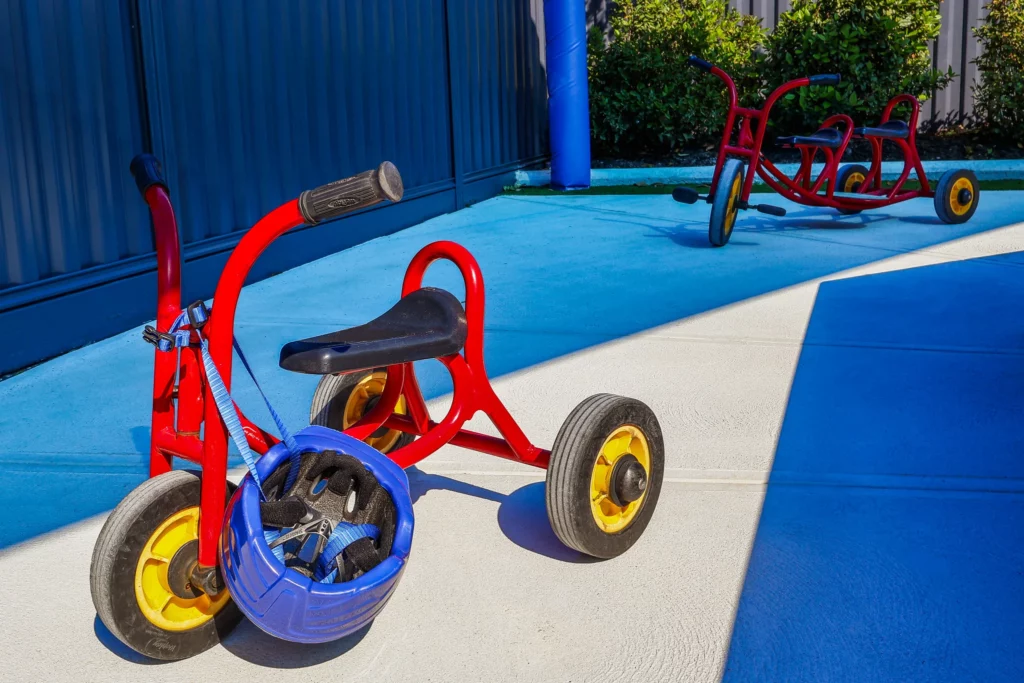 Bright red tricycles with yellow wheels parked on a sunlit concrete surface at Buttercups Perth Childcare. A blue helmet rests on one of the tricycles, highlighting the emphasis on safety. The area is partially shaded by a blue canopy, with greenery and a blue pole visible in the background, creating a vibrant and engaging environment for children.